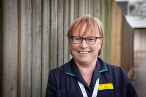 Photo of Hospice Matron, Liz Monaghan stood against a fence. She is smiling with her lanyard and name page on in nurses outfit