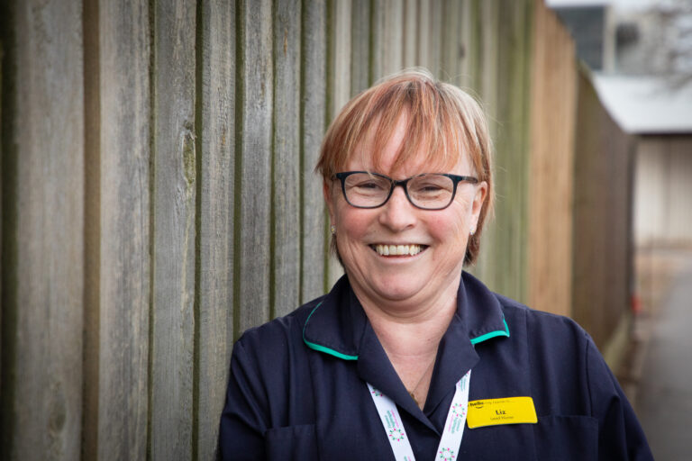 Photo of Hospice Matron, Liz Monaghan stood against a fence. She is smiling with her lanyard and name page on in nurses outfit